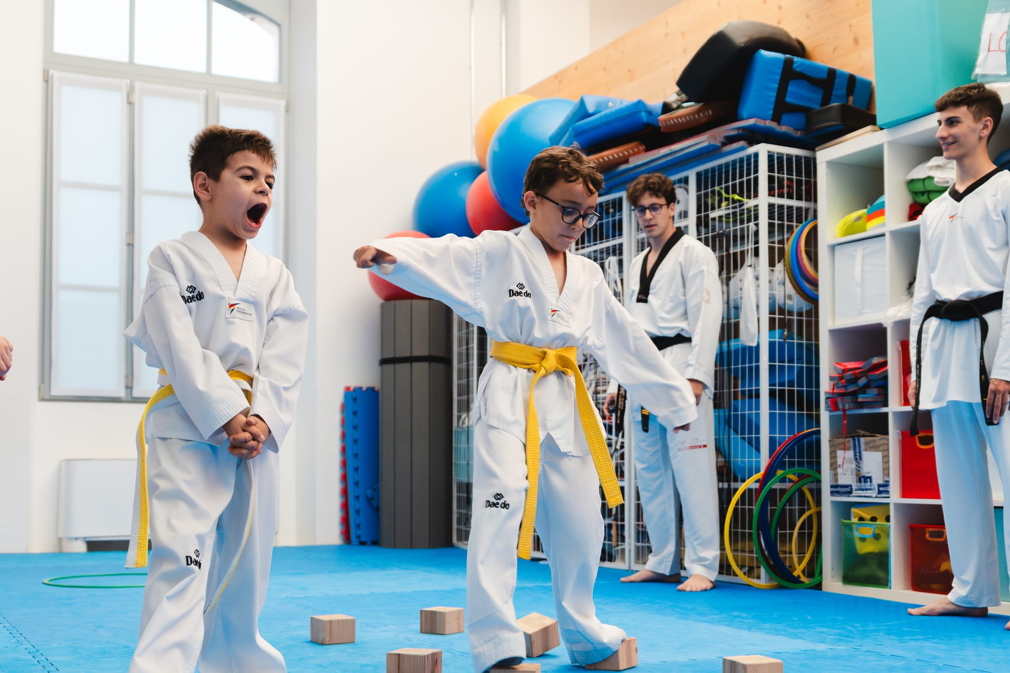 Due bambini con cintura gialla camminano in equilibrio sui blocchi di legno durante il percorso di coordinazione in palestra