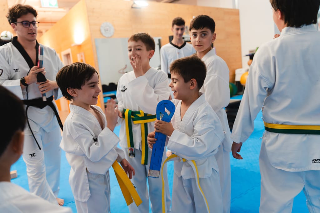 Bambini ridono insieme con le cinture gialle e verdi annodate in vita, uno tiene una paletta da allenamento